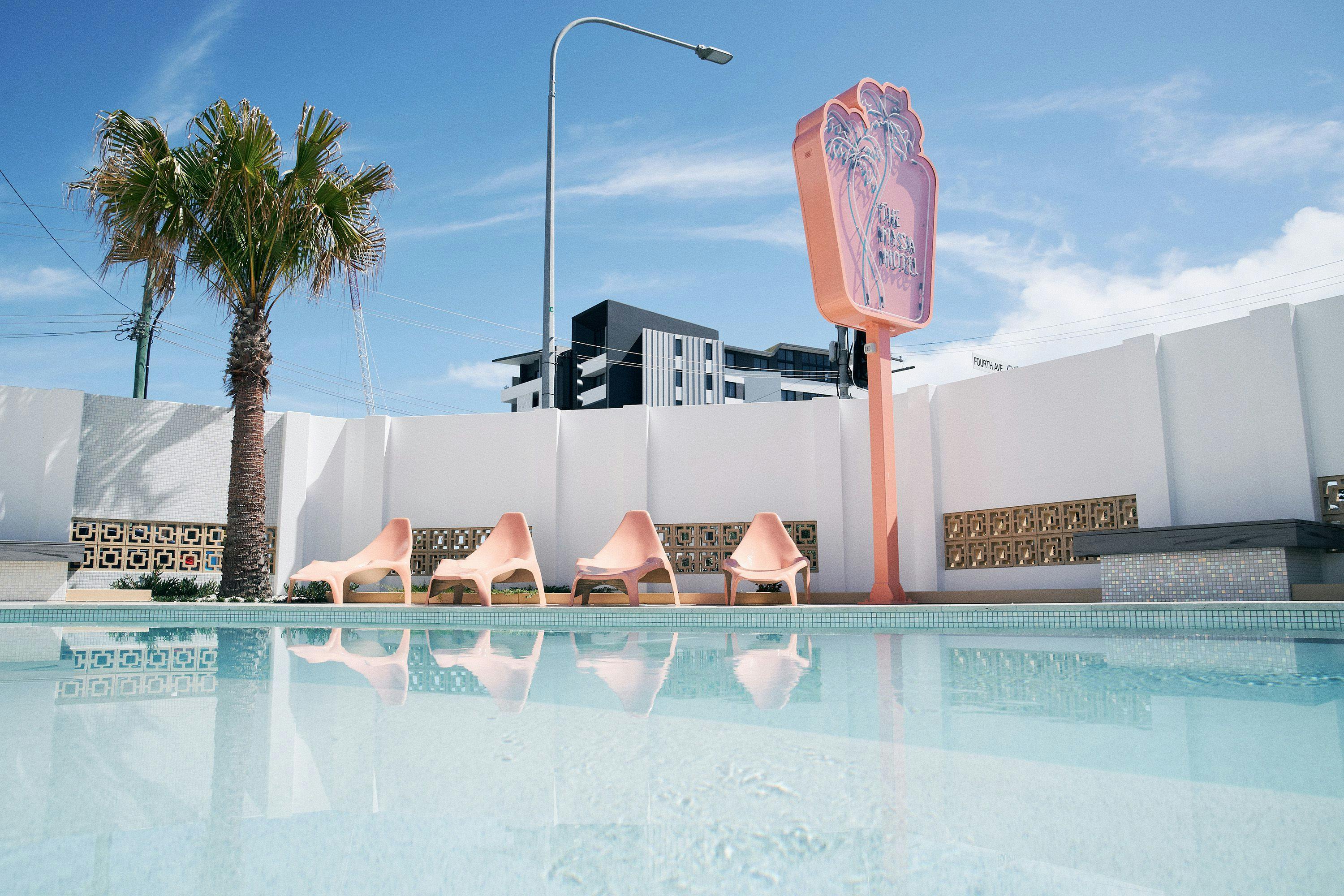 the exterior of a retro motel with kidney shaped pool and a neon pink sign and a palm tree